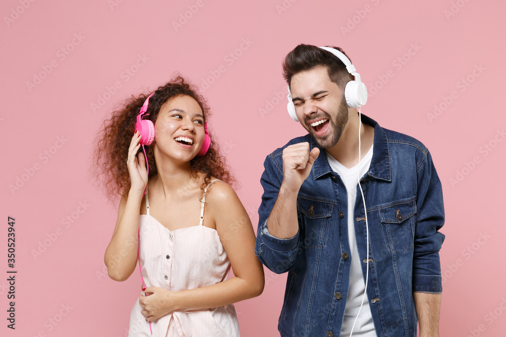Laughing young couple two friends european guy african american girl in casual clothes isolated on pastel pink background in studio. People lifestyle concept. Listen music with headphones, sing song. Laughing young couple two friends european guy african american girl in casual clothes isolated on pastel pink background in studio. People lifestyle concept. Listen music with headphones, sing song.