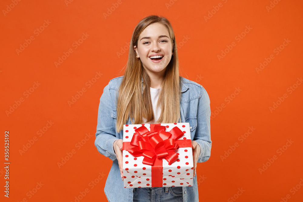 Laughing young woman girl in casual denim clothes isolated on orange background. Valentine's Day, Women's Day, birthday, holiday concept. Mock up copy space. Hold red present box with gift ribbon bow. Laughing young woman girl in casual denim clothes isolated on orange background. Valentine's Day, Women's Day, birthday, holiday concept. Mock up copy space. Hold red present box with gift ribbon bow.