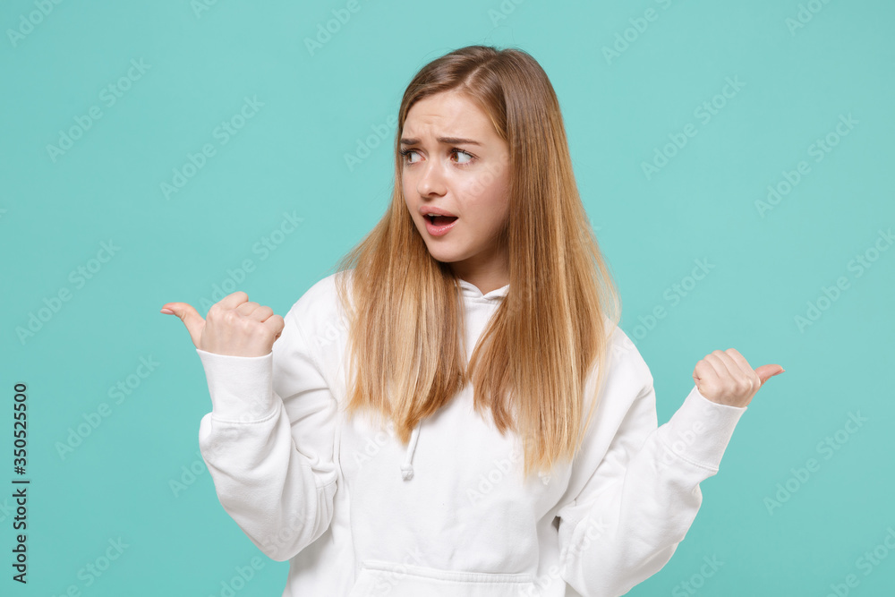 Perplexed young woman girl in casual white hoodie posing isolated on blue turquoise background studio portrait. People sincere emotions lifestyle concept. Mock up copy space. Pointing thumbs aside. Perplexed young woman girl in casual white hoodie posing isolated on blue turquoise background studio portrait. People sincere emotions lifestyle concept. Mock up copy space. Pointing thumbs aside.