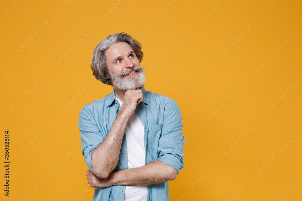 Pensive elderly gray-haired mustache bearded man in casual blue shirt posing isolated on yellow background studio. People lifestyle concept. Mock up copy space. Put hand prop up on chin, looking up. Pensive elderly gray-haired mustache bearded man in casual blue shirt posing isolated on yellow background studio. People lifestyle concept. Mock up copy space. Put hand prop up on chin, looking up.