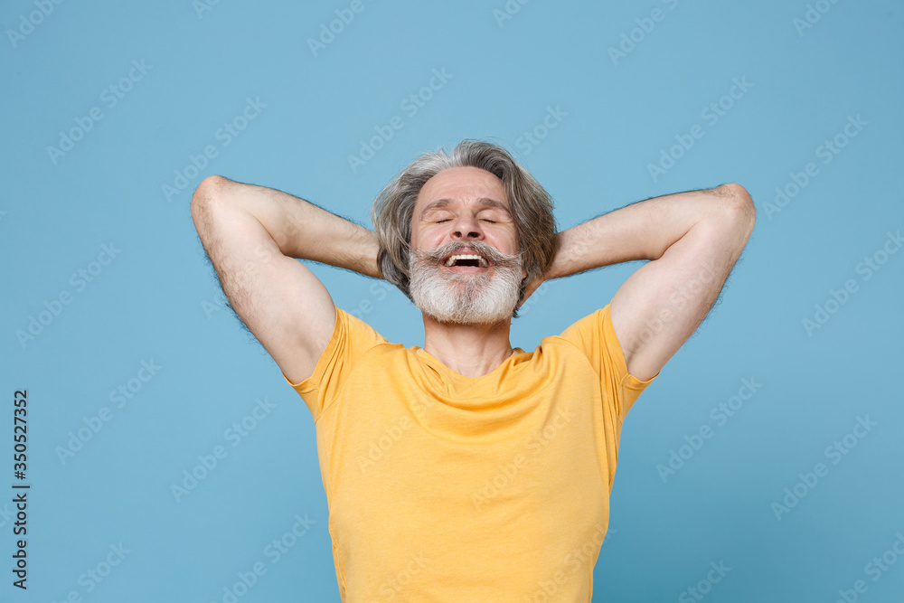 Relaxed elderly gray-haired mustache bearded man in casual yellow t-shirt isolated on blue background studio portrait. People lifestyle concept. Mock up copy space. Sleeping with hands behind head. Relaxed elderly gray-haired mustache bearded man in casual yellow t-shirt isolated on blue background studio portrait. People lifestyle concept. Mock up copy space. Sleeping with hands behind head.