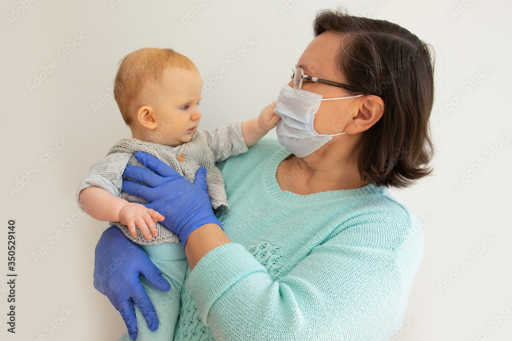 Grandma in mask and gloves holding baby granddaughter. Mature woman with little child posing isolated against white background. Pandemic and restriction concept