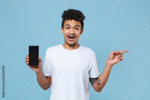 Excited young african american guy in white t-shirt isolated on blue background. People lifestyle concept. Mock up copy space. Hold mobile phone with blank empty screen, pointing index finger aside.