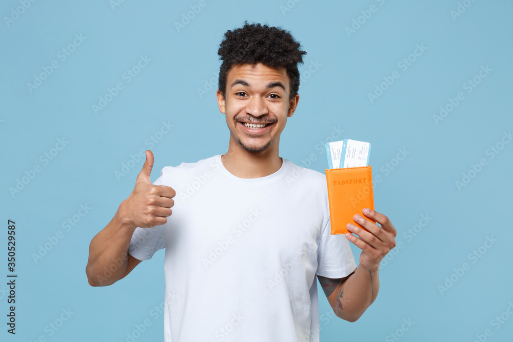 Smiling young african american guy in casual white t-shirt traveling abroad isolated on blue wall background. Air flight journey concept. Mock up copy space. Hold passport tickets, showing thumb up. Smiling young african american guy in casual white t-shirt traveling abroad isolated on blue wall background. Air flight journey concept. Mock up copy space. Hold passport tickets, showing thumb up.