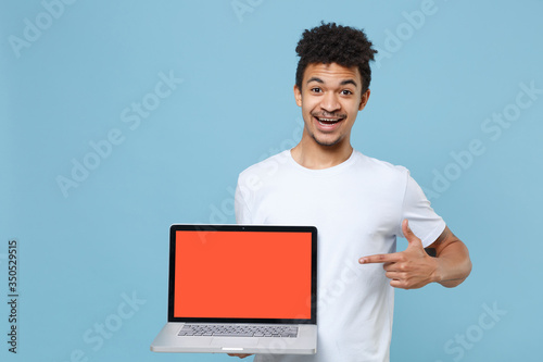 Funny young african american guy in casual white t-shirt isolated on blue background. People lifestyle concept. Mock up copy space. Pointing index finger on laptop pc computer with blank empty screen.