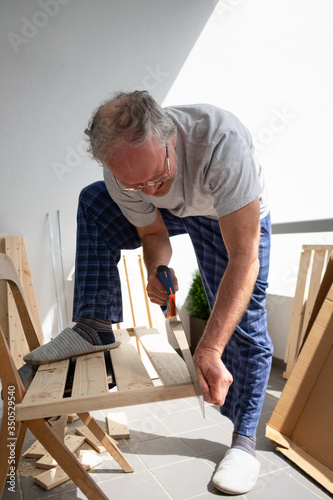 Senior grey-haired man in eyeglasses sawing wooden shelf and holding it with foot. Piece of wood laying on chair. House improving, DIY and home decoration during quarantine concept