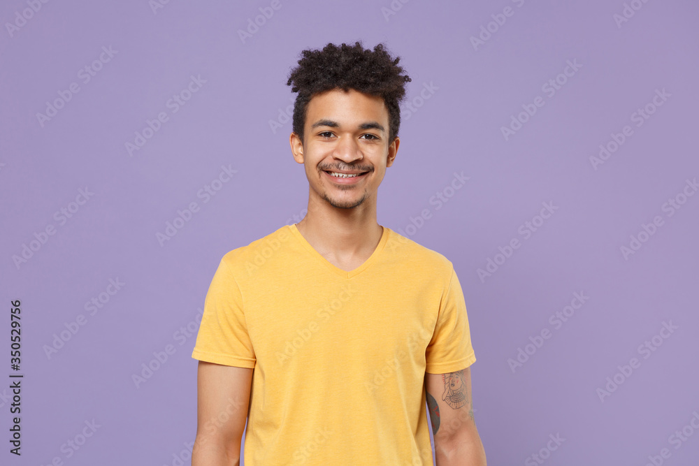 Smiling young african american guy in casual yellow t-shirt posing isolated on pastel violet background studio portrait. People sincere emotions, lifestyle concept. Mock up copy space. Looking camera. Smiling young african american guy in casual yellow t-shirt posing isolated on pastel violet background studio portrait. People sincere emotions, lifestyle concept. Mock up copy space. Looking camera.