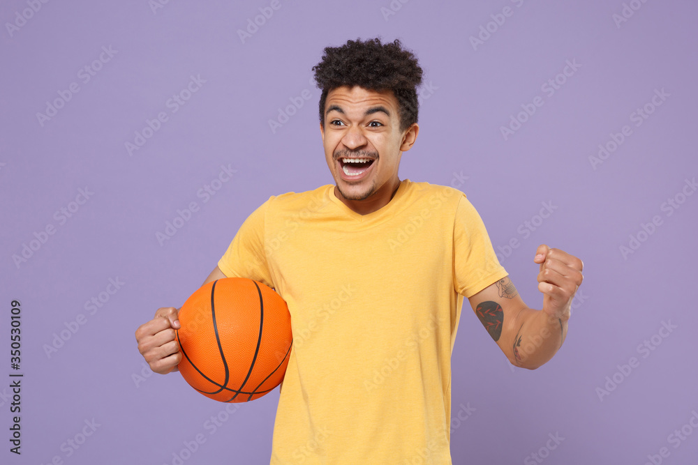 Excited young african american guy basketball player in yellow t-shirt isolated on pastel violet background. People emotions, sport leisure lifestyle concept. Play basketball hold ball clenching fist.
