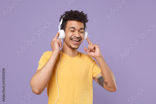 Funny young african american guy in casual yellow t-shirt posing isolated on pastel violet background. People lifestyle concept. Mock up copy space. Listen music with headphones keeping eyes closed.