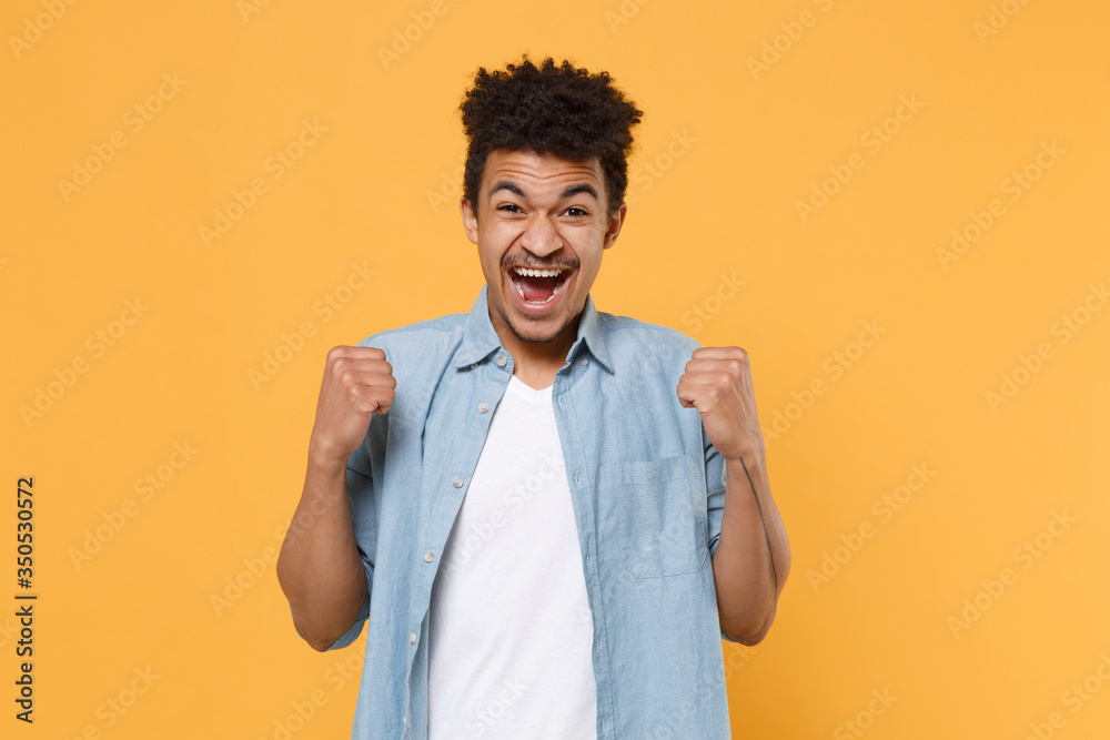 Joyful young african american guy in casual blue shirt posing isolated on yellow wall background studio portrait. People sincere emotions lifestyle concept. Mock up copy space. Doing winner gesture. Joyful young african american guy in casual blue shirt posing isolated on yellow wall background studio portrait. People sincere emotions lifestyle concept. Mock up copy space. Doing winner gesture.
