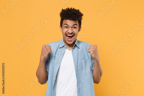 Joyful young african american guy in casual blue shirt posing isolated on yellow wall background studio portrait. People sincere emotions lifestyle concept. Mock up copy space. Doing winner gesture.