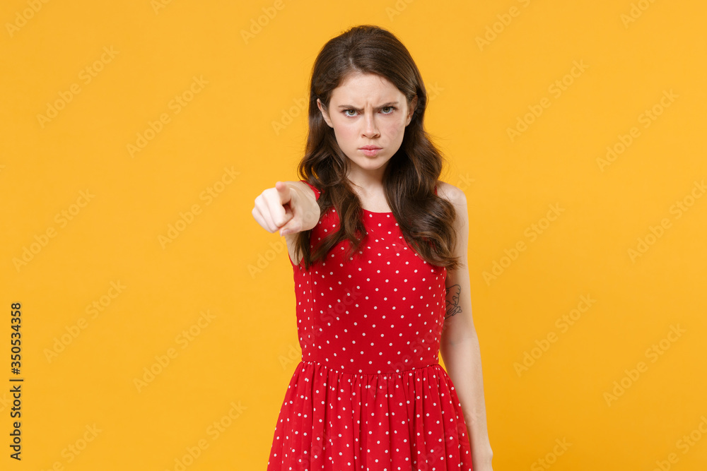 Displeased angry young brunette woman girl in red summer dress posing isolated on yellow wall background studio portrait. People lifestyle concept. Mock up copy space. Pointing index finger on camera. Displeased angry young brunette woman girl in red summer dress posing isolated on yellow wall background studio portrait. People lifestyle concept. Mock up copy space. Pointing index finger on camera.