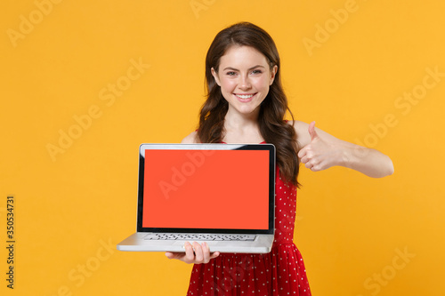 Smiling young brunette woman girl in red summer dress isolated on yellow background. People lifestyle concept. Mock up copy space. Hold laptop pc computer with blank empty screen, showing thumb up.