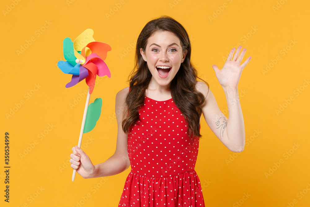 Excited young brunette woman girl in red summer dress posing isolated on yellow wall background studio. People lifestyle concept. Mock up copy space. Hold colorful pinwheel windmill, spreading hands. Excited young brunette woman girl in red summer dress posing isolated on yellow wall background studio. People lifestyle concept. Mock up copy space. Hold colorful pinwheel windmill, spreading hands.
