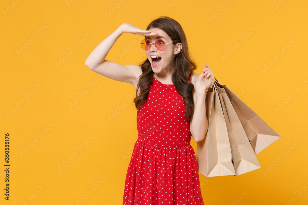 Excited girl in red summer dress, eyeglasses isolated on yellow background. People lifestyle concept. Hold package bag with purchases after shopping, hold hand at forehead looking far away distance.