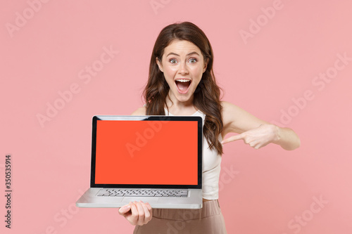 Excited young woman girl in light casual clothes isolated on pastel pink background. People lifestyle concept. Mock up copy space. Pointing index finger on laptop pc computer with blank empty screen. © ViDi Studio
