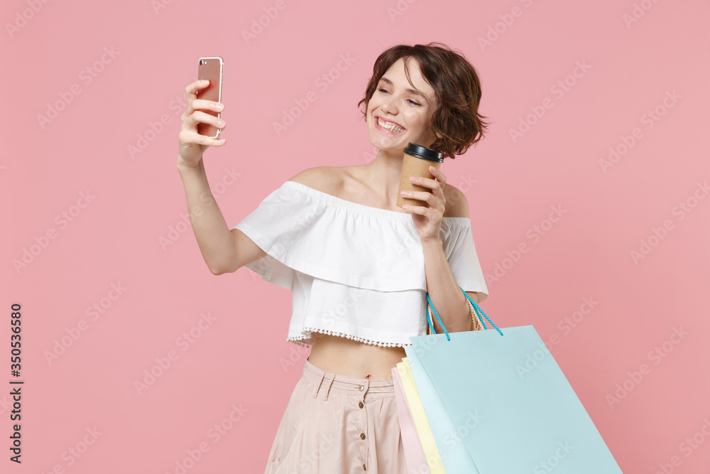 Smiling young woman girl in summer clothes isolated on pink background studio. Shopping discount sale concept. Hold package bag with purchases, cup of coffee or tea, doing selfie shot on mobile phone. Smiling young woman girl in summer clothes isolated on pink background studio. Shopping discount sale concept. Hold package bag with purchases, cup of coffee or tea, doing selfie shot on mobile phone.