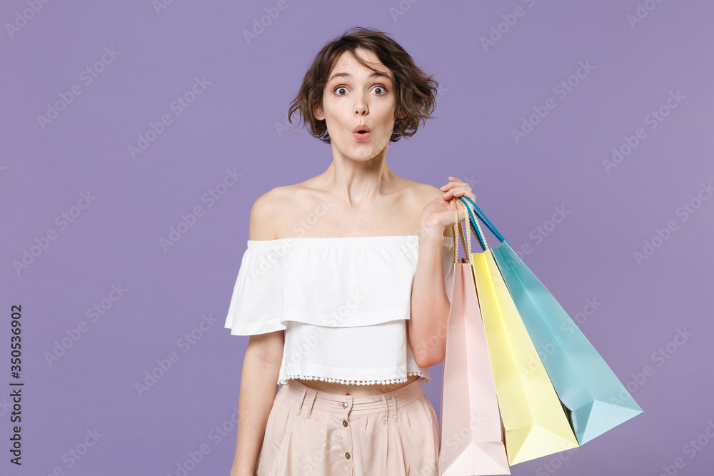 Shocked young woman girl in summer clothes hold package bag with purchases isolated on pastel violet background studio portrait. Shopping discount sale concept. Mock up copy space. Looking camera. Shocked young woman girl in summer clothes hold package bag with purchases isolated on pastel violet background studio portrait. Shopping discount sale concept. Mock up copy space. Looking camera.
