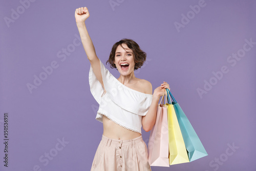 Happy young woman girl in summer clothes hold package bag with purchases isolated on pastel violet background in studio. Shopping discount sale concept. Mock up copy space. Clenching fist like winner.