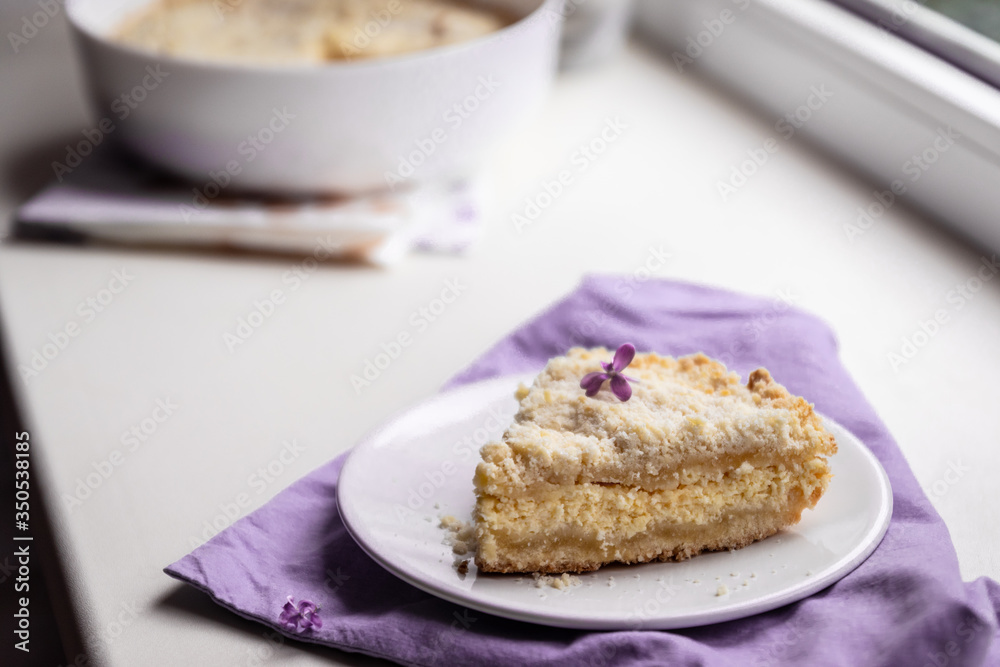 A piece of delicious cheesecake on a white plate on the windowsill, in the background a bouquet of lilacs
