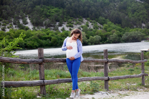 Adult pregnant woman relaxing in the nature against the lake . Pretty girl looking to the tummy  with love, female dressed blue jeans, white t-shirt, and shirt © Вероника Зеленина