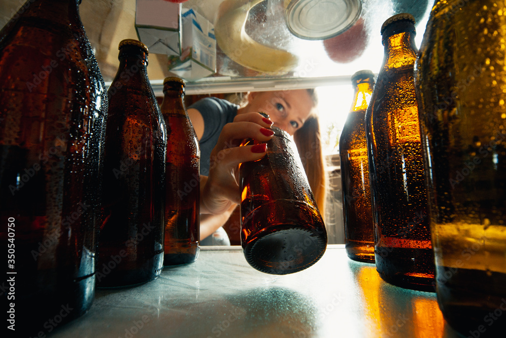 Caucasian woman takes cold refreshing beer from out the fridge, inside