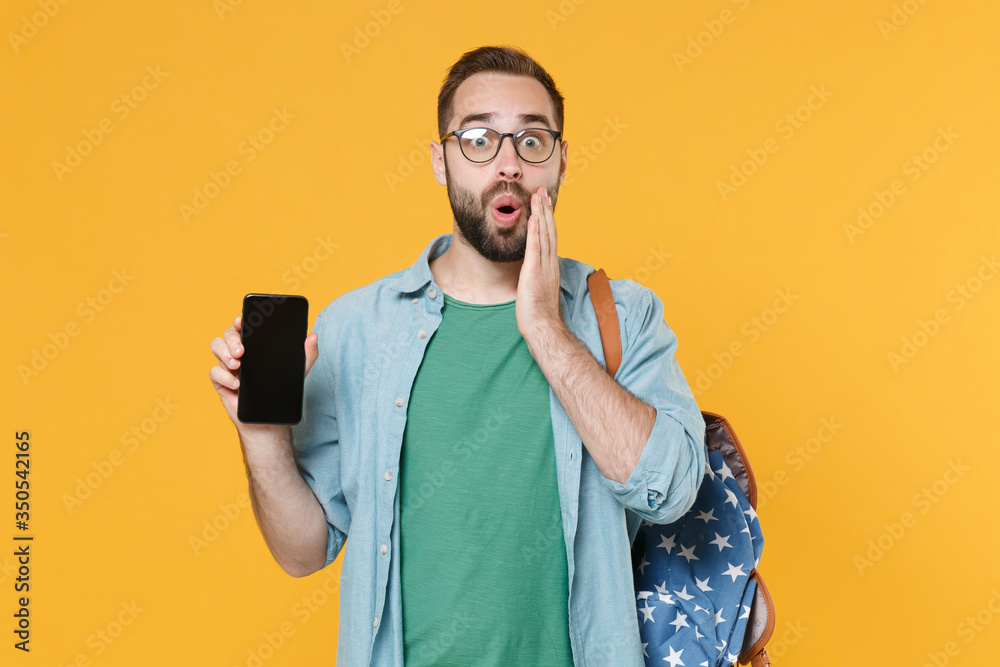 Shocked man student in casual clothes glasses backpack isolated on yellow background. Education in high school university college concept. Hold mobile phone with blank empty screen put hand on cheek. Shocked man student in casual clothes glasses backpack isolated on yellow background. Education in high school university college concept. Hold mobile phone with blank empty screen put hand on cheek.