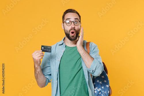 Shocked young man student in casual clothes glasses with backpack isolated on yellow background in studio. Education in high school university college concept. Hold credit bank card put hand on cheek.