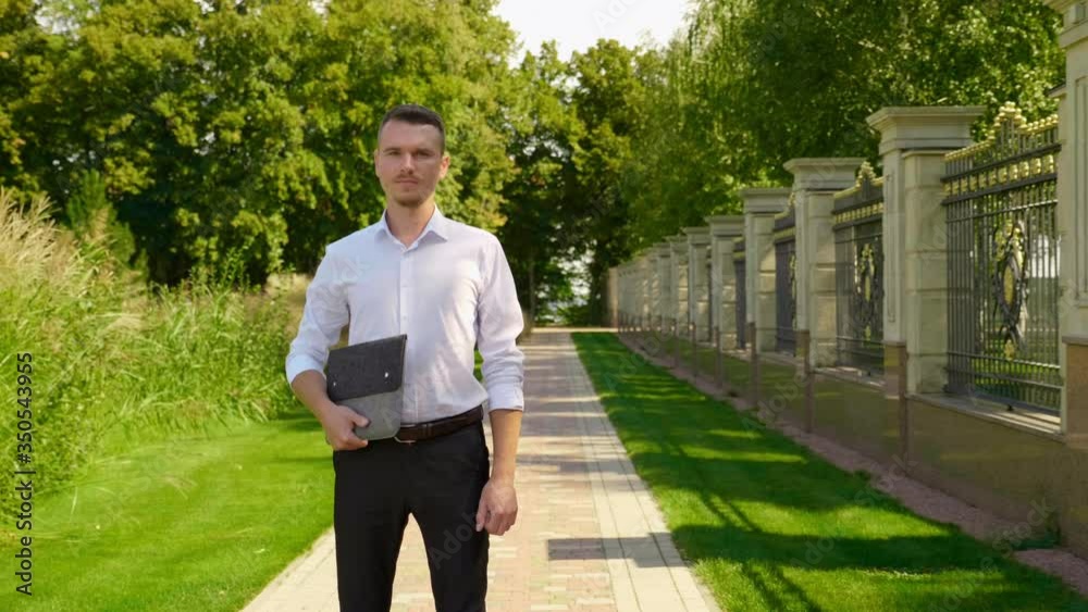 Businessman is standing in a park with laptop in his hands and looking at the camera. Freelancer man in a classic white shirt and trousers. 4K footage Businessman is standing in a park with laptop in his hands and looking at the camera. Freelancer man in a classic white shirt and trousers. 4K footage