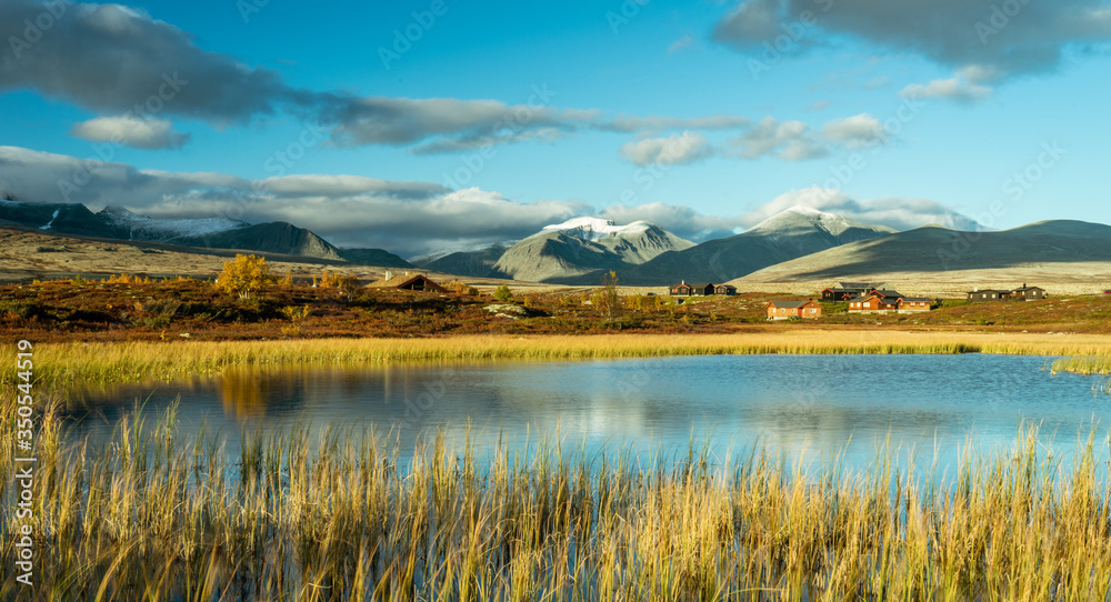 Rondane mountain chain covered with snow during golden hour and sunset. Cabins and lake covered in warm light. Mountains reflects in the lake. Autumn colors. Norway and travelling concept.