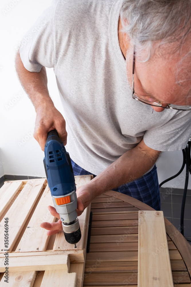 Concentrated man fixing shelf with electric screwdriver. Carpenter working at his apartment balcony with wood. House improving and home decoration during quarantine concept