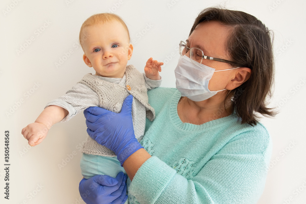 Happy grandma in medical mask and gloves holding cute baby granddaughter. Mature woman with little child posing isolated against white background. Epidemic and child protection concept