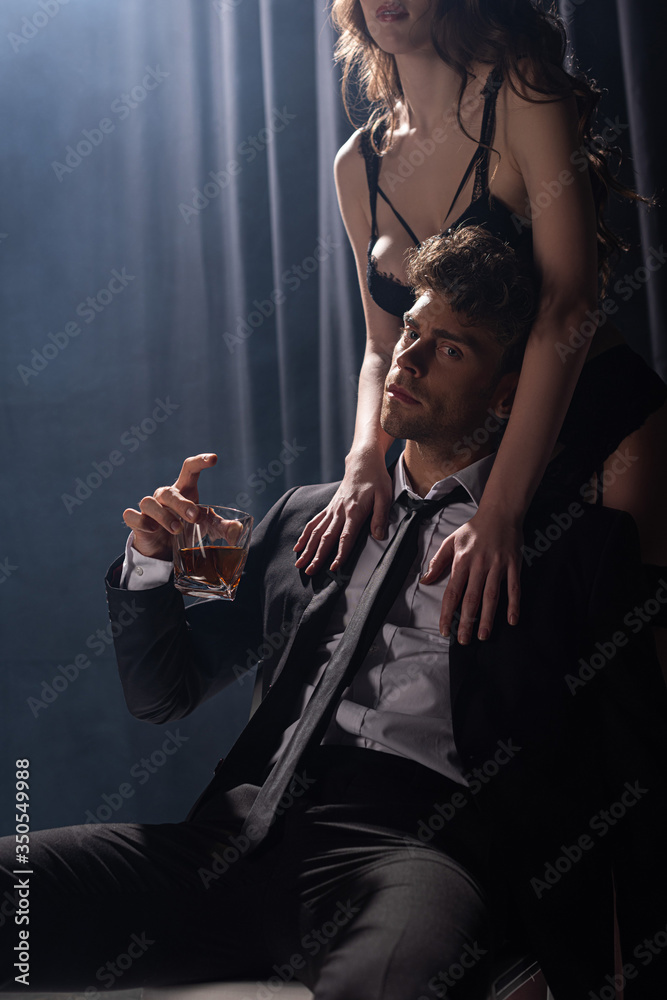 seductive woman touching handsome man sitting and holding glass of whiskey on black