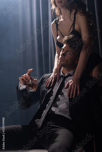 seductive woman touching handsome man sitting and holding glass of whiskey on black