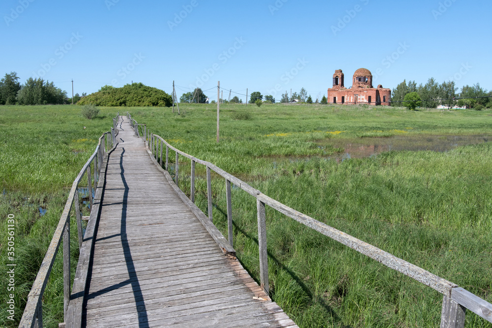 Wooden pathway over a swampy area between Staroye Rakomo and Novoye Rakomo villages and ruins of Znamenskaya church. Novgorod Oblast, Russia.