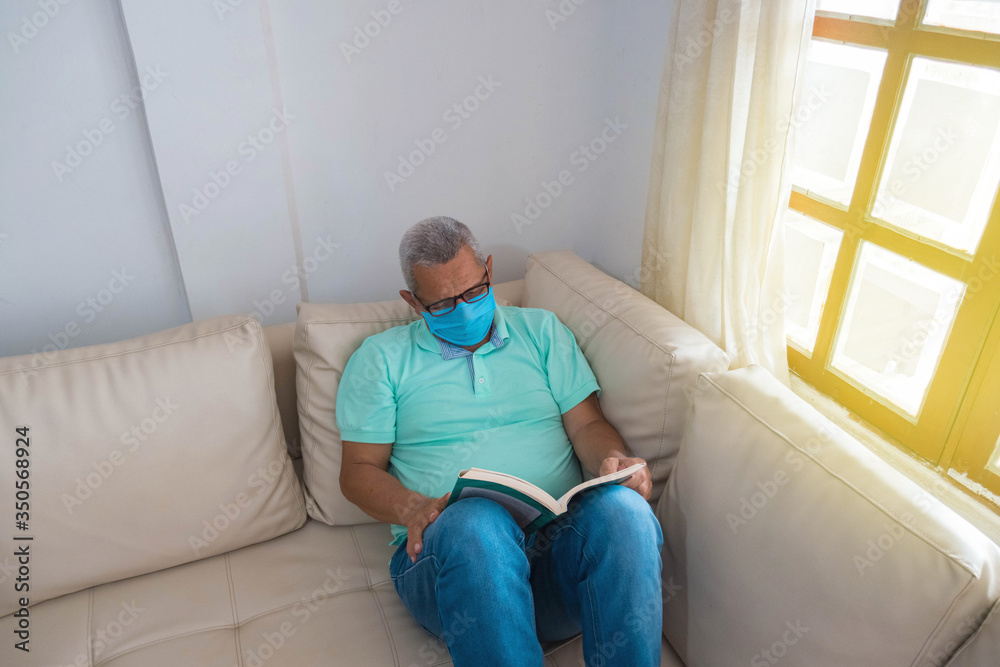 A retired man in a medical mask reading a book on his couch at home A retired man in a medical mask reading a book on his couch at home