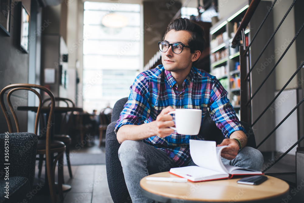 Positive young man in casual outfit with daily planner drinking coffee in cafe