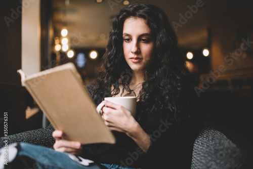 Thoughtful young brunette lady reading book with cup of hot drink