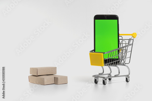 A shopping cart in an online store next to a smartphone on one white background. Paper boxes in a construction trolley
