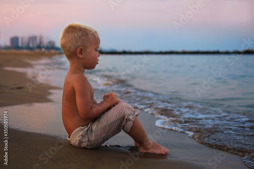 Little baby boy sitting on the beach near sea and looking on the waves, boy on the seaside in sunset