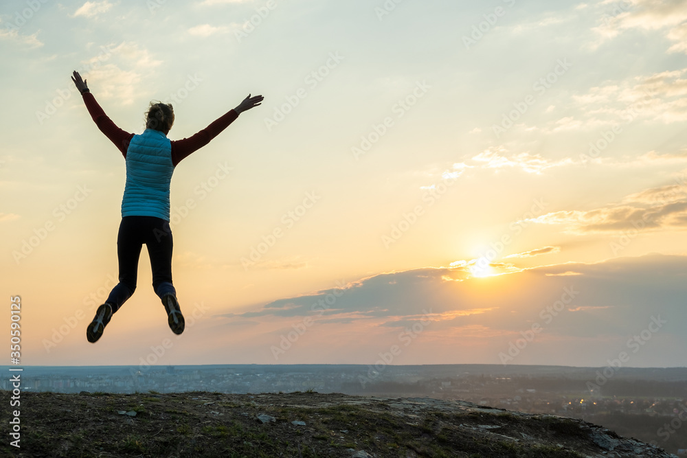 Silhouette of a woman hiker jumping alone on empty field at sunset in mountains. Female tourist raising her hands up in evening nature. Tourism, traveling and healthy lifestyle concept. Silhouette of a woman hiker jumping alone on empty field at sunset in mountains. Female tourist raising her hands up in evening nature. Tourism, traveling and healthy lifestyle concept.