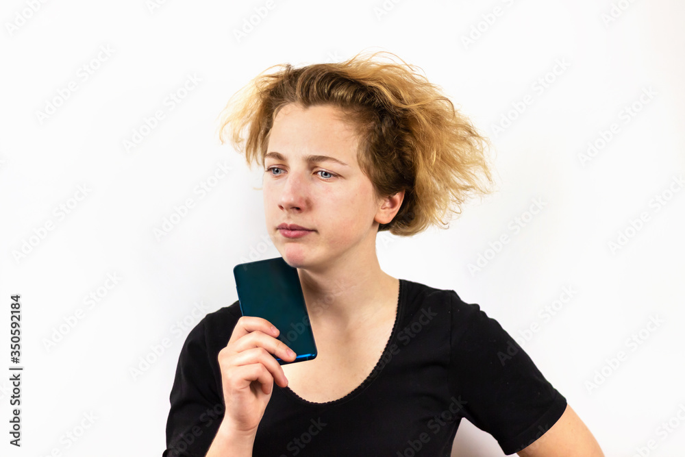 A girl with yellow hair holds a smartphone next to her face and looks down thoughtfully, sadly, against a white background. Modern technologies, youth, emotions. A girl with yellow hair holds a smartphone next to her face and looks down thoughtfully, sadly, against a white background. Modern technologies, youth, emotions.