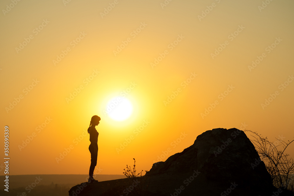 Silhouette of a woman hiker standing alone near big stone at sunset in mountains. Female tourist on high rock in evening nature. Tourism, traveling and healthy lifestyle concept.