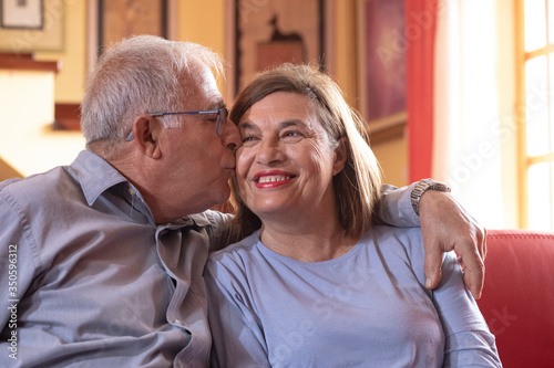 coppia di anziani si abbraccia felice e sorride nel divano del salotto di casa