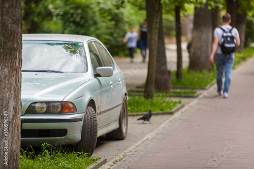 Car parked in pedestrian zone under trees along street with walking people on summer day.