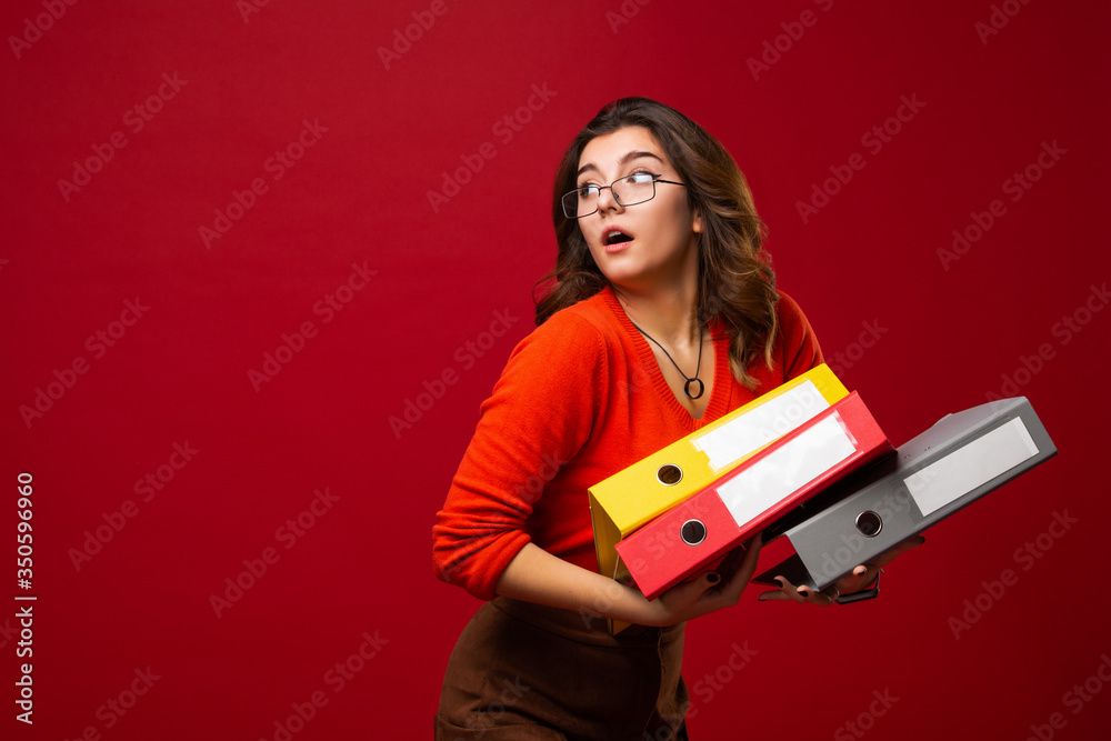 The girl in glasses carries folders, with a fright on her face on a red background. Mood stress, panic, accounting reporting, tax audit, exams The girl in glasses carries folders, with a fright on her face on a red background. Mood stress, panic, accounting reporting, tax audit, exams