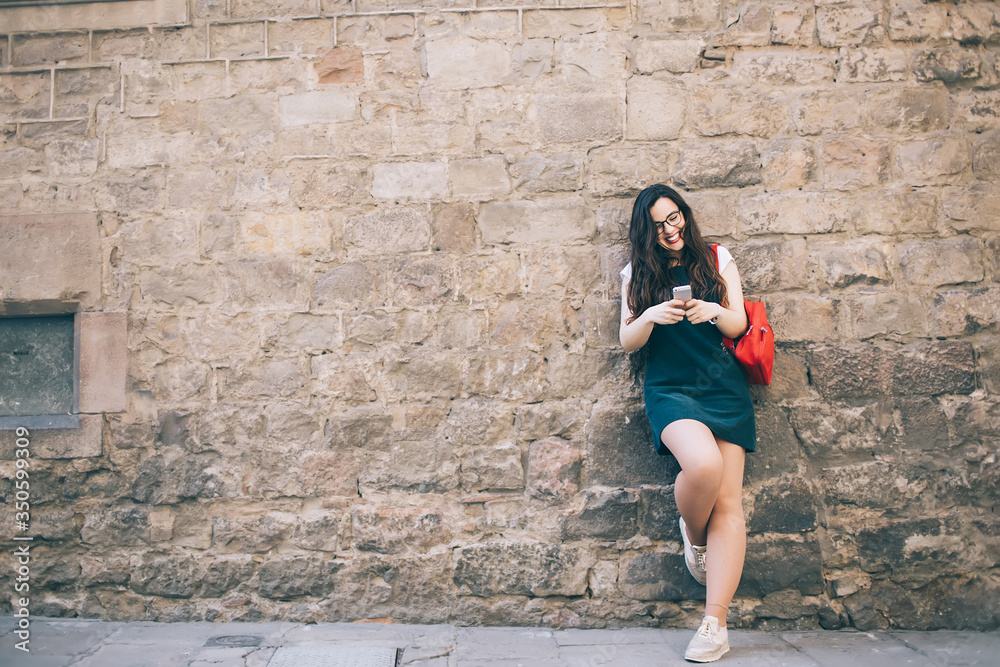 Cheerful caucasian female millennial satisfied with 4g connection to internet on mobile phone chatting online, smiling hipster girl in trendy wear share media in blog exploring city on journey