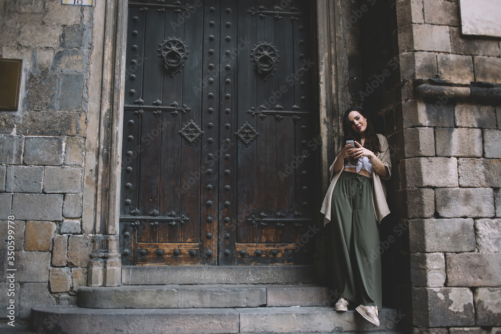 Full length portrait of caucasian female in trendy outfit standing near old building on street using mobile phone for blogging,attractive woman 20s dressed in stylish apparel checking mail on cellular
