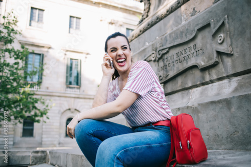 Emotional hipster girl in casual wear having funny conversation on mobile phone sitting on street, cheerful young woman 20s laughing while talking on smartphone enjoying tariffs and free time outdoors