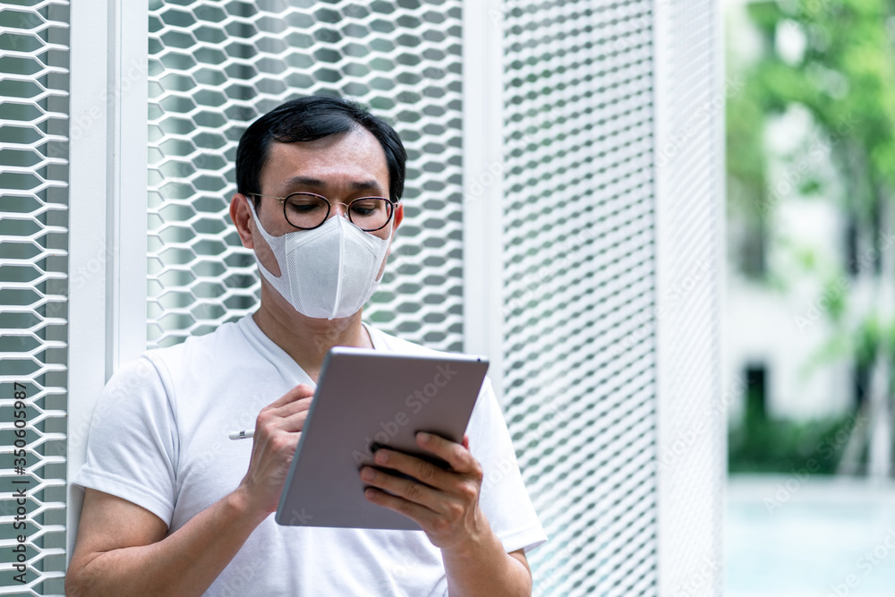 Man wearing a face pollution mask to protect himself from Covid-19. Asian male in casual T-shirts using tablet computer while relaxing outdoor. People working at home